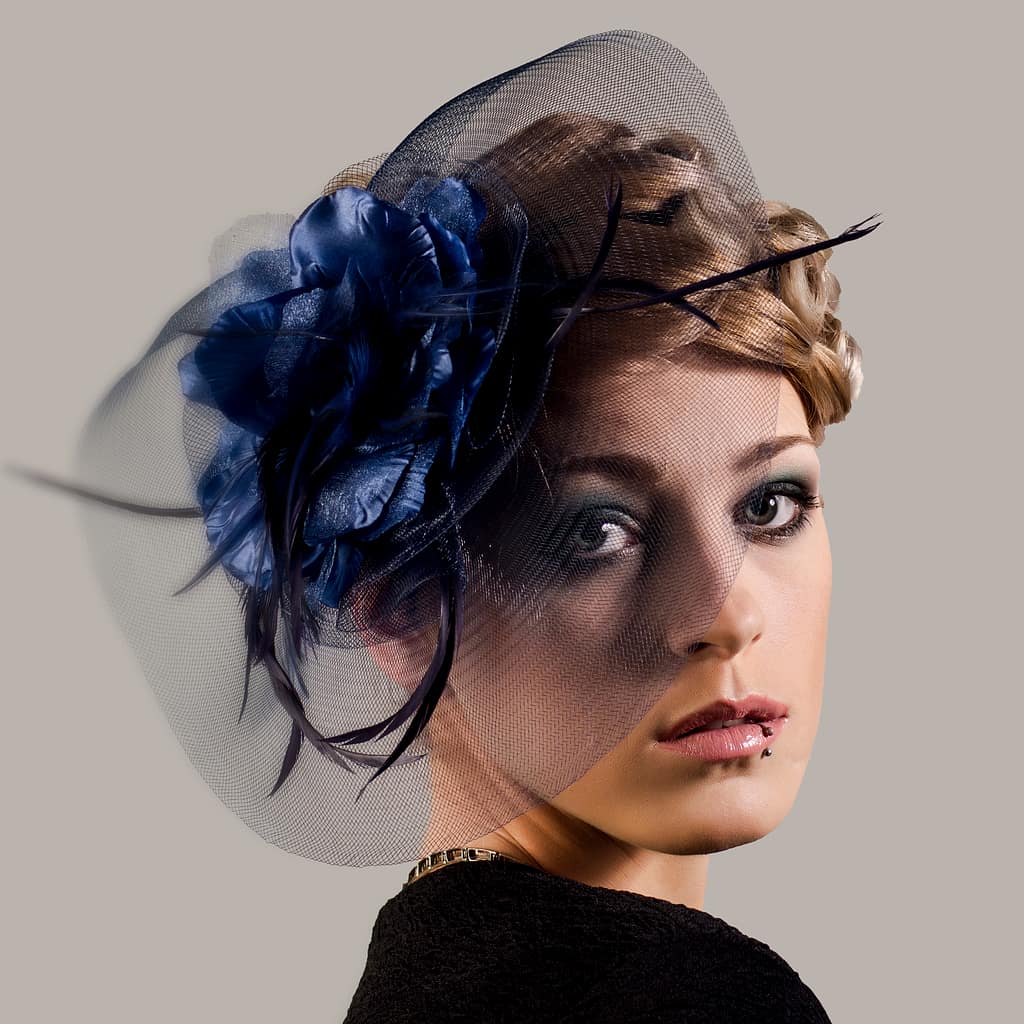 Jak przygotować się do portretowej sesji zdjęciowej w studiu i w plenerze? Kompleksowy przewodnik od fotografa 2 Artystyczny portret kobiety w eleganckim kapeluszu z granatową ozdobą i woalką, styl retro fashion portrait z subtelnym światłem studyjnym i neutralnym tłem podkreślającym detale stylizacji i makijażu