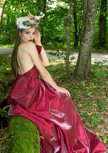 Modelka w czerwonej sukni – sesja bajkowa w lesie Modelka w czerwonej sukni siedząca na pniu drzewa podczas sesji zdjęciowej w lesie