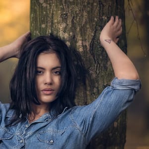 Portret przy drzewie Portret kobiety w koszuli jeansowej opierającej się o drzewo, fotografia plenerowa beauty z naturalnym światłem, subtelny klimat lifestyle