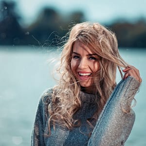 Lifestyle nad wodą Uśmiechnięta kobieta nad jeziorem w swetrze, naturalna sesja lifestyle outdoor z rozwianymi włosami, portret w chłodnej kolorystyce i plenerowym klimacie