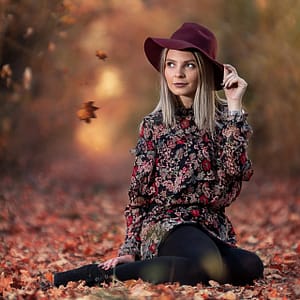 Jesień – portret lifestyle Jesienna fotografia portretowa kobiety siedzącej na liściach w bordowym kapeluszu, naturalne światło, ciepłe kolory jesieni, klimat lifestyle i fashion outdoor, profesjonalna sesja zdjęciowa plenerowa