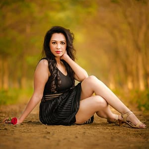 Czarna sukienka w jesiennym lesie Portret kobiety siedzącej na leśnej drodze w czarnej sukience z czerwonym kwiatem, artystyczna fotografia plenerowa glamour z ciepłym światłem jesiennego lasu i płytką głębią ostrości podkreślającą sylwetkę